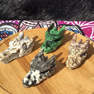 May include: Four carved dragon head sculptures made of different types of stone. The sculptures are displayed on a wooden surface. The dragon heads are carved in a realistic style and have intricate details. The stones used are green, white, black, and brown.