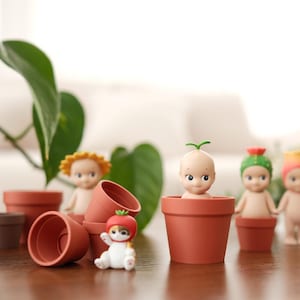 Puede incluir: Una colección de pequeñas figuras decorativas en macetas de terracota. Las figuras están desnudas con varios tocados, incluyendo un brote, un tomate y una flor. La escena está ambientada en una mesa de madera con plantas en el fondo.