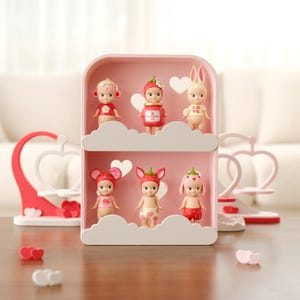 Może przedstawiać: Różowa półka z półkami w kształcie chmur zawiera sześć małych figurek w różnych kostiumach o tematyce owocowej. Półka stoi na drewnianym stole, z dekoracjami w kształcie serca i białą sofą w tle.