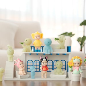 Può includere: Una collezione di figurine colorate, tra cui figure con disegni floreali, angelici e a tema animale, disposte su una struttura bianca con accenti blu. Alcune figure sono in piccoli vasi. La scena è ambientata su un tavolo di legno.