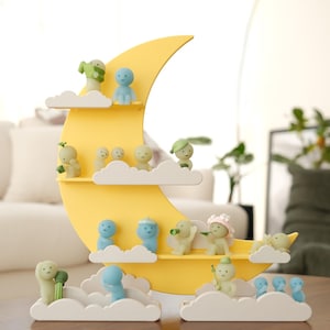 May include: A whimsical yellow crescent moon shelf with cloud-shaped platforms, displaying small figurines. The figurines are in various poses and colors, including blue and green. The shelf is on a wooden table.