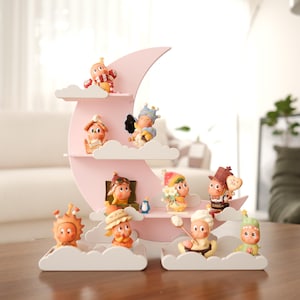 May include: A pink crescent moon-shaped shelf with cloud-shaped platforms, displaying whimsical figurines. The figurines are dressed in various outfits and poses. The shelf sits on a wooden table, creating a charming display.