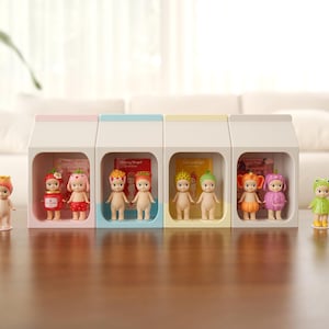 Op de afbeelding: Een verzameling kleine, poppenachtige beeldjes, elk met unieke outfits en accessoires, tentoongesteld in individuele witte, melkpakvormige vitrines. De beeldjes staan op een donkerbruine tafel, met een witte bank op de achtergrond.
