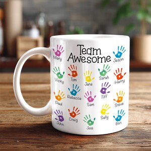 May include: A white ceramic mug featuring the black text Team Awesome at the top, surrounded by multiple colorful handprint illustrations in shades of red, blue, green, yellow, orange, and purple, each labeled with a name underneath.
