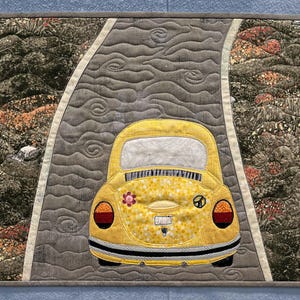 May include: A quilted wall hanging depicting a yellow Volkswagen Beetle on a grey road. The car has a flower and peace sign on the back. The road is bordered by textured, colourful fabric, and the piece is framed in grey.