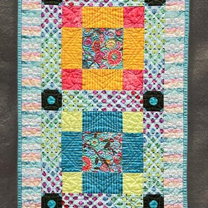 May include: A colorful quilted table runner with a floral and geometric pattern. The runner features squares of yellow, pink, and teal fabric with floral and striped designs. The edges are bordered with a light blue fabric. The name "Laura Kushner" is visible.
