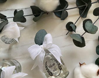 May include: A clear glass bottle with a white ribbon and a black label that says "Welcome Baby" surrounded by greenery and cotton balls.
