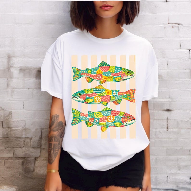 May include: A white short-sleeved t-shirt featuring a graphic print of three fish stacked vertically. Each fish is decorated with a colorful patchwork pattern of hearts, flowers, and geometric shapes against a background of vertical light orange stripes.