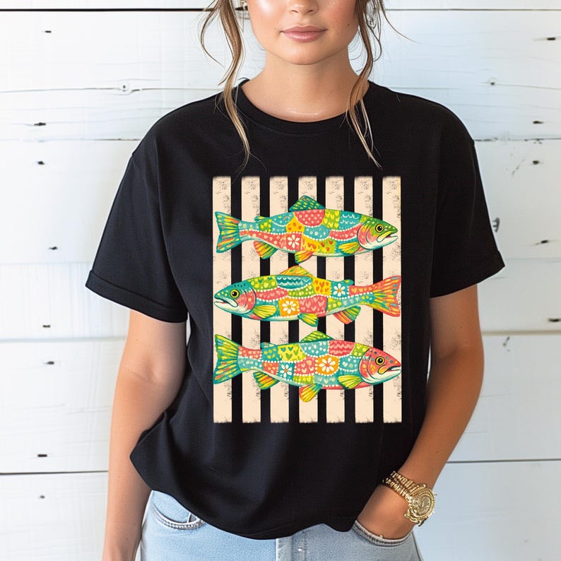 May include: A black short-sleeved t-shirt featuring a central graphic of three fish stacked vertically. Each fish is decorated with a colorful, multi-patterned design including floral and geometric shapes, set against a background of vertical black stripes.