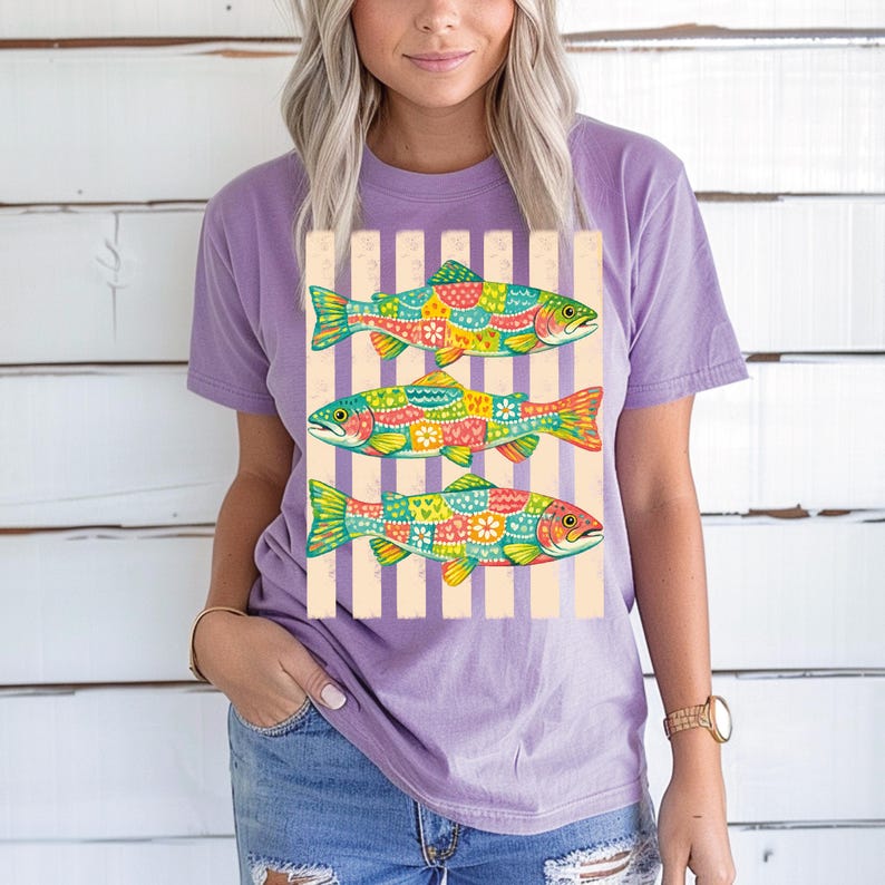 May include: A light purple short-sleeve t-shirt featuring a graphic print of three fish stacked vertically. Each fish is decorated with a colorful patchwork pattern of hearts, flowers, and geometric shapes against a background of vertical stripes.