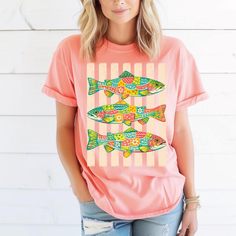 May include: A coral-colored short-sleeve t-shirt featuring a graphic print of three fish stacked vertically. Each fish is decorated with a colorful patchwork pattern of flowers, hearts, and geometric shapes against a background of vertical stripes.