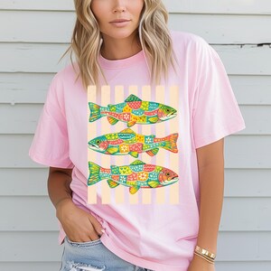 May include: A light pink t-shirt featuring a graphic print of three fish stacked vertically. Each fish is decorated with a colorful, intricate pattern of hearts, flowers, and dots in shades of green, yellow, orange, red, and blue against a striped background.