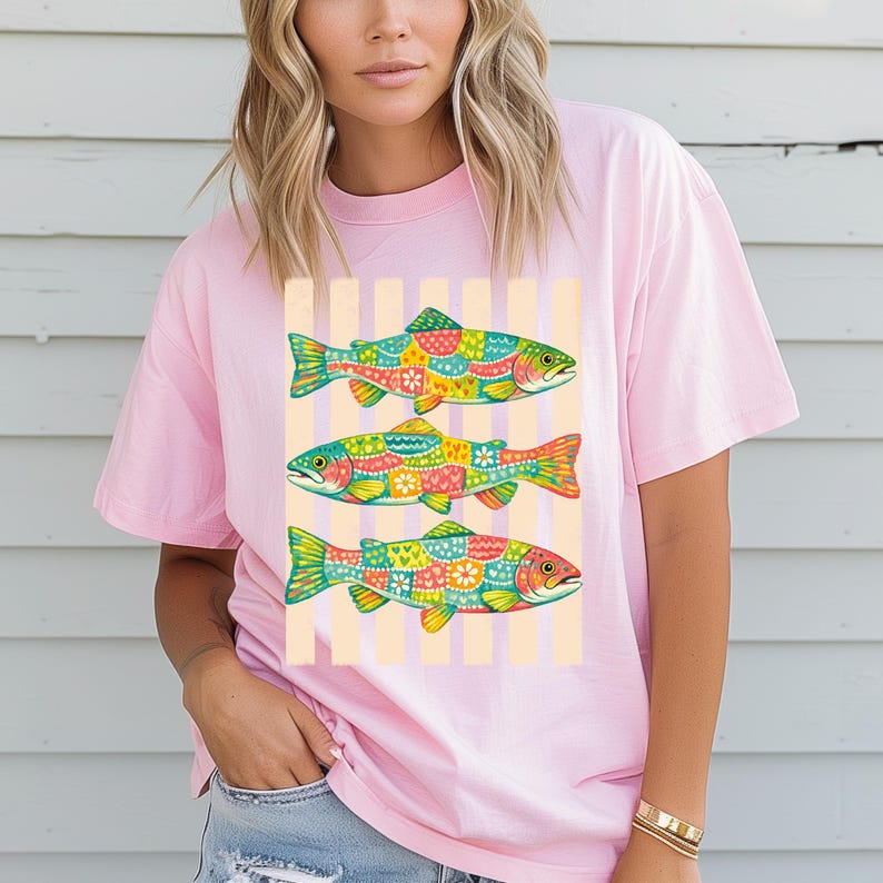 May include: A light pink t-shirt featuring a graphic print of three fish stacked vertically. Each fish is decorated with a colorful, intricate pattern of hearts, flowers, and dots in shades of green, yellow, orange, red, and blue against a striped background.