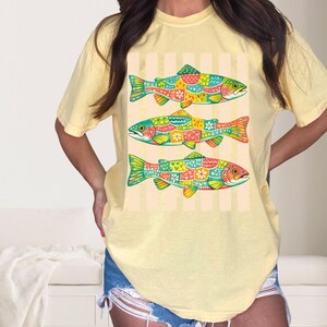 May include: A pale yellow short-sleeve t-shirt featuring a graphic print of three fish stacked vertically. Each fish is decorated with a colorful patchwork pattern of hearts, flowers, and geometric shapes against a background of vertical stripes.