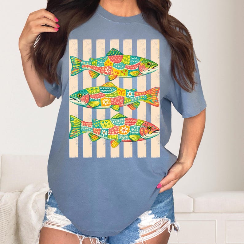 May include: A dusty blue short-sleeve t-shirt featuring a graphic print of three fish stacked vertically. Each fish is decorated with a colorful patchwork pattern of hearts, flowers, and dots, set against a background of vertical beige stripes.