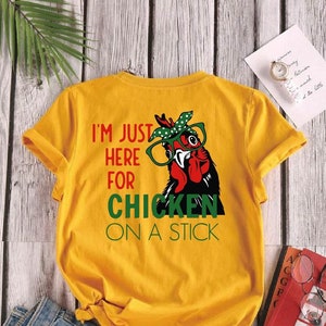 May include: Yellow short-sleeved t-shirt with a graphic of a chicken wearing glasses and a bandana. The text on the shirt reads "I'm just here for chicken on a stick."