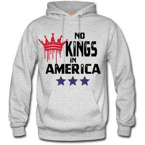 May include: A light gray hoodie with a front pocket and a hood. The front of the hoodie features the text "NO KINGS IN AMERICA" with a red crown graphic and three blue stars.