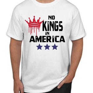 May include: White t-shirt with the text "NO KINGS IN AMERICA" in black, with a red crown graphic above. Three blue stars are below the text. The shirt is a crew neck.