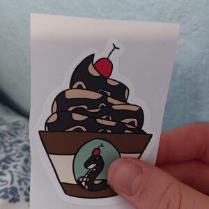 May include: A sticker of a black and brown snake shaped like a sundae with a red cherry on top. The snake is curled up in a bowl with a white rim and a brown base. There is a smaller snake with a red cherry on top in a green circle on the side of the bowl.