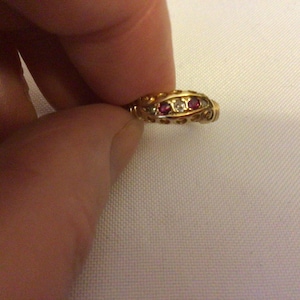 May include: A gold ring with three gemstones. The ring features two red gemstones and one clear gemstone. The ring is held by a person's fingers against a white background. The ring has an ornate design.