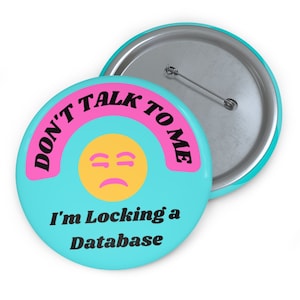Könnte beinhalten: Ein türkisfarbener Knopf mit einer pinkfarbenen und schwarzen Grafik, auf der "Sprich mich nicht an, ich sperre eine Datenbank" steht. Die Grafik enthält auch ein gelbes Emoji mit einem traurigen Gesicht.