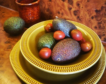 May include: A green ceramic bowl with a decorative rim, filled with avocados and red tomatoes. The bowl is on a matching green plate, and there is a brown ceramic mug in the background.