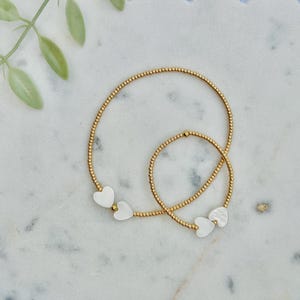 May include: Two gold-toned beaded bracelets featuring small, white, heart-shaped charms. Each bracelet is constructed from tiny, uniform metallic beads strung together, with two flat, white heart accents positioned near the clasp area of each piece.