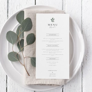 May include: A white menu card with a green leaf design and the word "MENU" in a simple font. The menu lists "Starters", "Main Course", and "Dessert" with a few food items listed under each category. The card is on a white plate with a sprig of eucalyptus leaves and a linen napkin.