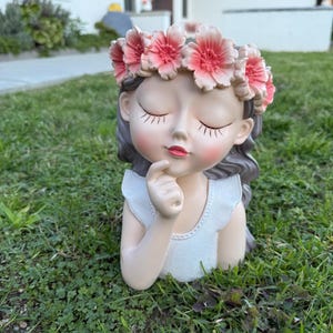 May include: A decorative planter shaped like a girl with closed eyes, wearing a flower crown. The planter is painted in soft, natural tones with a white top. The girl's hair is gray, and the flowers are pink.
