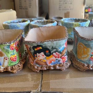 May include: Hand-painted ceramic planters with unique designs. One features a cat with strawberries, another a girl, and the third a cat with a hat. The planters have a textured, brown base and a light-colored interior.