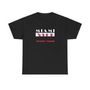 May include: Black t-shirt with the words "MIAMI VICE STUNT TEAM" in white, red, and blue. The text is arranged in a rectangular design on the front of the shirt. The shirt is made of a soft, comfortable material.