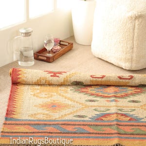 May include: A handwoven rug with a geometric pattern in shades of beige, red, orange, and blue. The rug has a fringe border and is made of natural fibers.