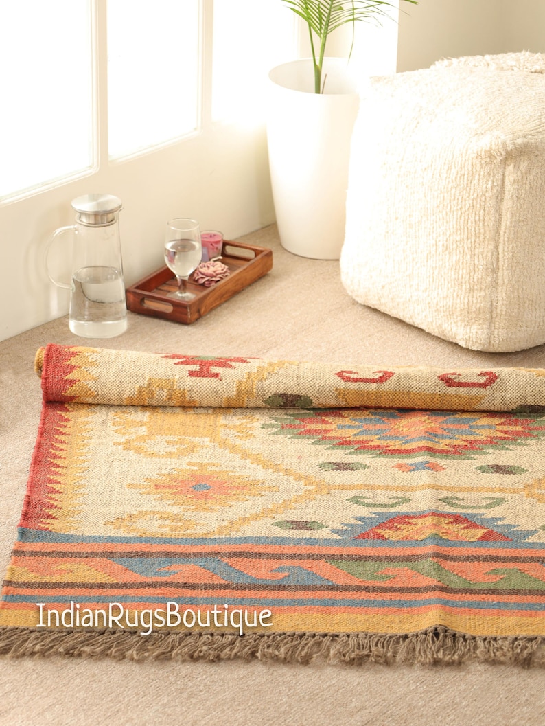 May include: A handwoven rug with a geometric pattern in shades of beige, red, orange, and blue. The rug has a fringe border and is made of natural fibers.