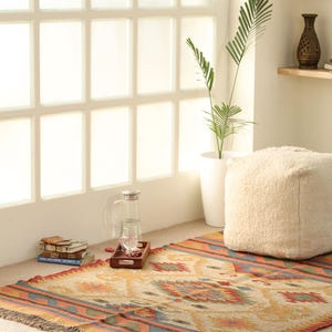 May include: A colorful, handwoven rug with a geometric pattern in shades of orange, red, blue, and green. The rug has a fringe border and is laid out on a light brown floor. A white pouf sits on the rug, and a plant in a white pot is visible in the background.