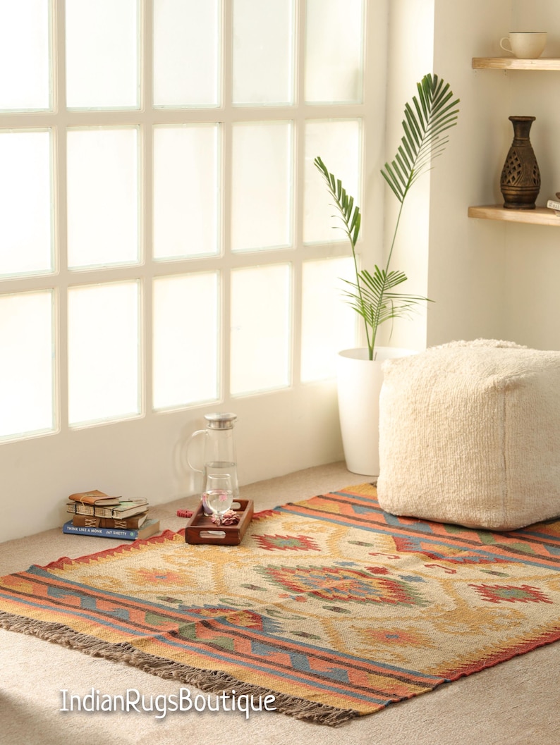 May include: A colorful, handwoven rug with a geometric pattern in shades of orange, red, blue, and green. The rug has a fringe border and is laid out on a light brown floor. A white pouf sits on the rug, and a plant in a white pot is visible in the background.