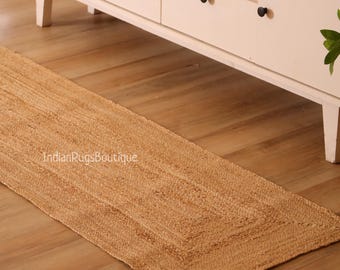 Puede incluir: Una alfombra de pasillo rectangular de yute tejido en color beige natural. La alfombra presenta un diseño con bordes y está colocada sobre un suelo de madera. El texto "IndianRugsBoutique" es visible en la alfombra.