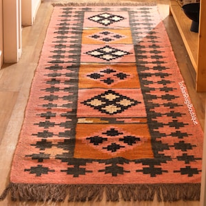 May include: A rectangular, hand-woven rug with a geometric design. The rug features a central panel of orange and pink, with diamond and cross patterns in black and white. The edges are pink with a repeating black cross pattern and a fringed border.