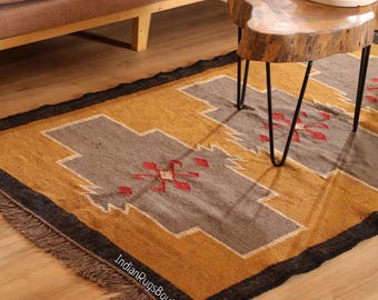 May include: A rectangular rug with a geometric pattern in shades of gold, gray, and red. The rug has a black border and fringed edges. The text "IndianRugsBoutique" is visible on the rug.