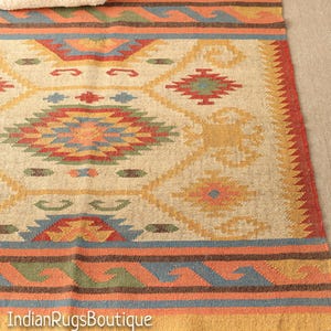 May include: A beige, brown, red, blue, and green patterned rug with fringe. The rug has a geometric design with a central diamond shape.