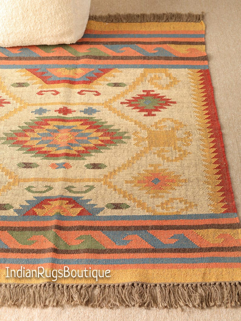 May include: A beige, brown, red, blue, and green patterned rug with fringe. The rug has a geometric design with a central diamond shape.