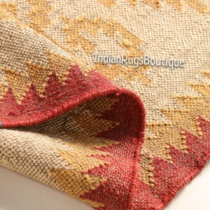 May include: Close-up of a woven rug with a geometric pattern in shades of beige, red, and yellow. The rug has a fringed edge and is folded in half. The text "IndianRugsBoutique" is visible in the image.