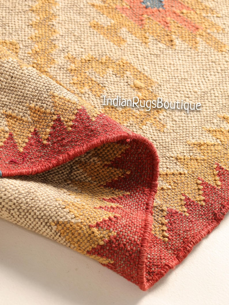May include: Close-up of a woven rug with a geometric pattern in shades of beige, red, and yellow. The rug has a fringed edge and is folded in half. The text "IndianRugsBoutique" is visible in the image.