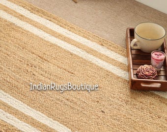 May include: A natural jute runner rug with two off-white stripes. The rug is rectangular and has a woven texture. The text "IndianRugsBoutique" is printed on the rug. A wooden tray with a teacup, candle, and rose is on the right.