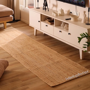 Könnte beinhalten: Ein rechteckiger Jute-Läuferteppich in Naturtönen. Der Teppich liegt auf einem Holzboden, mit einem weißen Schrank und Dekoration im Hintergrund. Die Textur des Teppichs ist sichtbar, und die Gesamtästhetik ist neutral und erdig.
