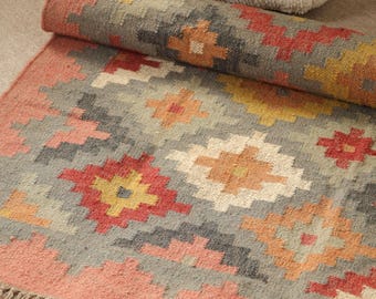 May include: A rolled-up, rectangular rug with a geometric pattern in shades of gray, red, yellow, and peach. The rug has a peach border and fringe along one edge. The text "IndianRugsBoutique" is visible on the rug.
