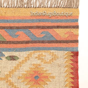 May include: A handwoven rug with a geometric pattern in shades of beige, yellow, orange, red, blue, and green. The rug has a fringe border and the text "IndianRugsBoutique" is visible in the top center of the rug.