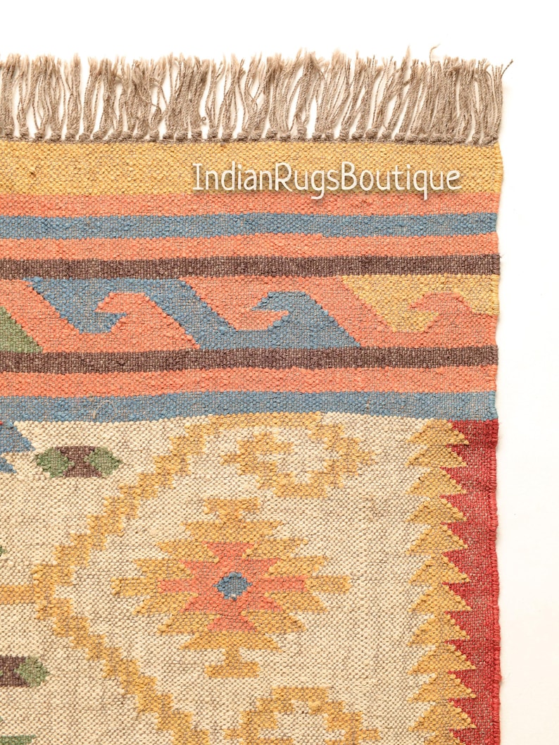 May include: A handwoven rug with a geometric pattern in shades of beige, yellow, orange, red, blue, and green. The rug has a fringe border and the text "IndianRugsBoutique" is visible in the top center of the rug.