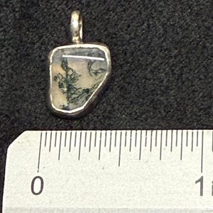 May include: A pendant featuring a translucent, irregularly shaped stone with dark green moss-like inclusions and a thin white horizontal line, encased in a silver-toned metal bezel with a top bail, positioned above a ruler showing 1 inch.