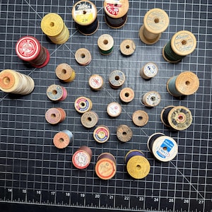 May include: A collection of vintage wooden spools of thread in various colors, including red, yellow, green, and blue. Some spools have labels with text like "CLARK'S" and "DUTY". The spools are arranged on a black grid surface.