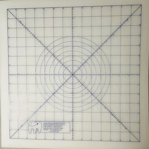 May include: A white, gridded design mat with concentric circles and lines. The mat is marked with numbers and has a blue grid. The mat is designed for use with a rotary cutter and ruler. The mat is made by Polar Notions and has the company's logo and contact information printed in the bottom right corner.